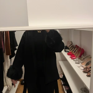 Svart kappa med fuskpälsdetaljer - Stilren svart kappa med bred krage och bälte i midjan. Ärmarna har snygga svarta fuskpälsdetaljer som ger jackan en lyxig vibe. Perfekt för dig som vill sticka ut med en trendig och elegant look.