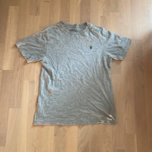 Grå t-shirt från Polo Ralph Lauren - Klassisk grå t-shirt från Polo Ralph Lauren med liten broderad logga på bröstet. T-shirten är i mjuk bomull och har rund hals och korta ärmar. Perfekt basic-plagg med clean stil och diskret branding.