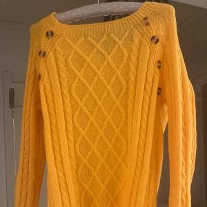 Gul stickad tröja med knappar - NY - Säljer en snygg gul stickad tröja med flätstickat mönster och dekorativa knappar längs axlarna. Tröjan har rund halsringning och långa ärmar, perfekt för att liva upp garderoben med lite färg.