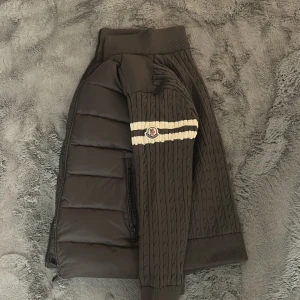 Moncler mörkgrå puffer med stickade ärmar - Snygg mörkgrå pufferjacka från Moncler med stickade ärmar och rygg i kabelstickat mönster. Jackan har två vita ränder och Moncler-logga på ärmen, dragkedja framtill och ribbade muddar. Perfekt för dig som vill ha en unik och sportig look.