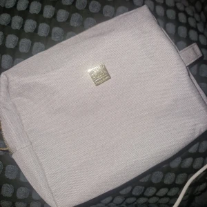 Necessär  - Stilren beige necessär från Upper House Gothia Towers. Tillverkad i tåligt tyg med guldfärgad logga framtill och dragkedja upptill. Insidan har flera praktiska fack och är fodrad i glansigt material. Perfekt för att organisera dina småsaker. Finns tecken på användning på insidan, men fyller sin funktion.