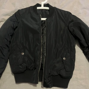 Svart bomberjacka med dragkedja - Snygg svart bomberjacka med klassisk ribbad mudd i ärmslut och midja. Jackan har silverfärgad dragkedja framtill, fickor med tryckknappar och en extra ficka med dragkedja på ena ärmen. Tillverkad i glansig polyester för en stilren look.