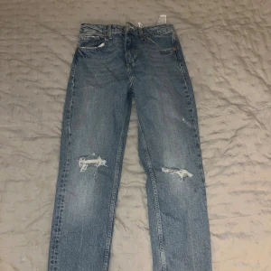 Blå ripped jeans från Zara, strl 36 - Snygga blå jeans från Zara i storlek 36 med raka ben och slitna detaljer på båda knäna. Klassisk femficksmodell med normal passform och hög midja. Perfekta för en avslappnad och trendig look.