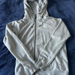 Grå Nike hoodie med dragkedja XS - Säljer en stilren grå hoodie från Nike i storlek XS. Tröjan har hel dragkedja, huva med snörning och två fickor framtill. Perfekt för chill dagar eller när du vill ha en sportig look. Tillverkad i mjuk bomullsblandning för extra komfort.