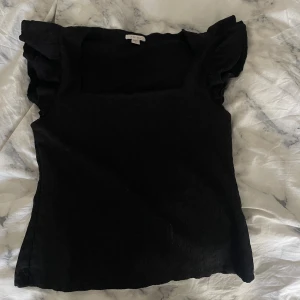 Svart volangtopp från Amisu - Säljer en svart topp från Amisu med fyrkantig ringning och volangdetaljer vid axlarna. Toppen är ärmlös och har en stilren, enkel design som passar perfekt till jeans eller kjol. Materialet känns mjukt och lätt mot huden.