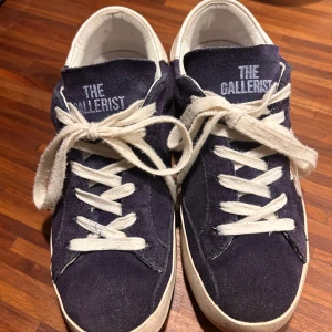 The Gallerist blå mockasneakers - Snygga sneakers från The Gallerist i mörkblå mocka med vita detaljer och snörning. Skorna har vit sula, vit klöverdetalj på sidan och blått klövermönster baktill. Tungan och hälen pryds av The Gallerist-loggan. Perfekta för dig som gillar unik stil.