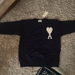 Mörkblå stickad tröja från Ami Paris - Säljer en mörkblå stickad tröja från Ami med ett vitt hjärta och bokstaven A på bröstet. Tröjan har rund hals, långa ärmar och breda ribbade muddar vid ärmslut och nederkant. Perfekt för dig som gillar stilrena detaljer och snygg design.