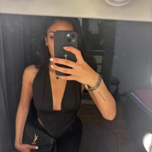 Svart omlott halterneck topp - Säljer en svart halterneck topp med djup v-ringning och omlottknytning i ryggen. Toppen är croppad med spetsig nederkant framtill och öppen rygg. OBS! Den har en liten defekt som knappt syns. Ni ser en inzomning på sista bilden.