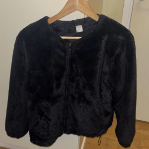 Svart teddyjacka från Lindex - Mysig svart teddyjacka från Lindex med dragkedja framtill och två stora fickor. Jackan har rund halsringning och är helfodrad, perfekt för kyliga dagar. Den har en loose passform och är tillverkad i mjukt syntetmaterial.