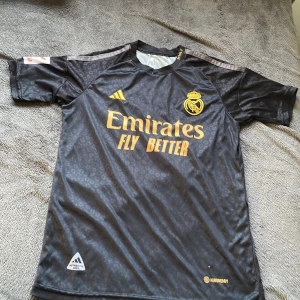 Real Madrid Mbappé svart matchtröja - Svart Real Madrid fotbollströja från Adidas med Mbappé och nummer 10 på ryggen. Tröjan har guldfärgade detaljer, klubbmärke och sponsortryck på bröstet samt La Liga-märke på ärmen. Materialet är glansigt polyester med subtilt leopardmönster och klassiska Adidas-ränder på axlarna. 