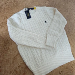 Vit kabelstickad tröja Polo Ralph Lauren - Kabelstickad vit tröja från Polo Ralph Lauren med klassisk rund halsringning och broderad marinblå logga på bröstet. Tröjan är i mjuk ull och har ribbade muddar vid ärmslut och nederkant. Perfekt för lager-på-lager under kyliga dagar.