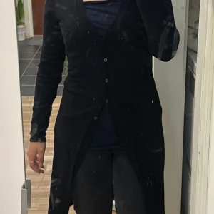 Svart lång kofta med knappar - Säljer en svart lång kofta med knappar framtill. Koftan har en smickrande v-ringning och långa ärmar. Perfekt att slänga över en t-shirt eller topp för en chill och stilren look.