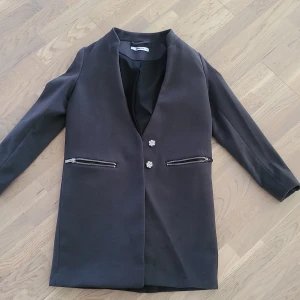 Svart kavaj från Gina Tricot - Stilren svart kavaj från Gina Tricot med raka axlar och dekorativa blommiga knappar framtill. Två fickor med dragkedja ger en cool touch. Perfekt för dig som vill ha en enkel men snygg look. Tunnare material och helfodrad.