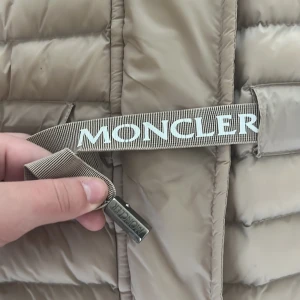 Beige Moncler dunjacka med huva - Snygg beige dunjacka från Moncler med huva och justerbart bälte i midjan. Jackan har broderad logga på ärmen och klassisk quiltad design. Perfekt för kyliga dagar och riktigt stilren look. S-M
