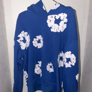 Denim tears hoodie - Säljer en blå hoodie med stora vita blommönster över hela tröjan. Tröjan har huva, magficka och långa ärmar. Perfekt för dig som gillar färg och vill sticka ut lite extra.