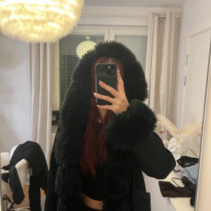 Svart pälsjacka med stor huva - Säljer en svart pälsjacka med oversized passform och stor fluffig huva. Jackan har breda pälsdetaljer längs huva, ärmslut och framkant. Perfekt för dig som vill sticka ut och hålla dig varm under vintern. Snygg och trendig look.