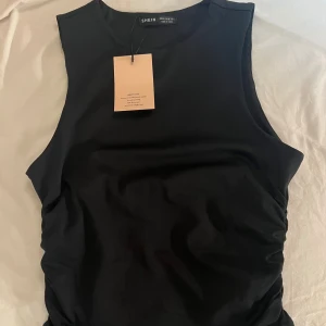 Svart topp - Svart, tight och ärmlös topp från SHEIN med rynkade sidor som ger en snygg detalj. Toppen har rund halsringning och är i ett stretchigt, mjukt material som sitter skönt mot kroppen. Sitter som s.