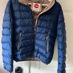 Blå pufferjacka från Colmar, M - Säljer en snygg blå pufferjacka från Colmar med beige insida och huva. Jackan har två dragkedjeförsedda fickor framtill och en cool logga på ärmen. Perfekt för kyliga dagar och riktigt skön att bära. Passar dig som gillar stilren och sportig look. Pris kan diskuteras 