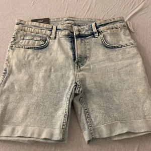Skinny fit jeansshorts ljus denim - Säljer ett par skinny fit jeansshorts i ljusblå tvättad denim. Klassisk femficksmodell med bälteshällor och dragkedja. Snyggt slitna detaljer och uppvikta benslut ger en modern vibe. Perfekta för dig som gillar en tightare passform och streetstil.