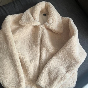 Beige teddyjacka Gina Tricot med dragkedja - Supermjuk teddyjacka från Gina Tricot beige färg. Jackan har en bred krage, lång ärm och stängs med dragkedja framtill. Perfekt för kyliga dagar och ger en mysig vibe till din outfit.
