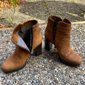 Bruna mocka stövletter från Ekonika - Snygga bruna stövletter i mocka från Ekonika med hög klack och rund tå. Skorna har dragkedja på sidan, dekorativt spänne och mjukt grått foder. Perfekta för höst och vinter när du vill ha både stil och komfort.