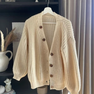 Beige stickad kofta med knappar - Mysig beige stickad kofta med grov struktur och stora bruna knappar framtill. Koftan har lång ärm, djup v-ringning och ribbade muddar vid ärmslut och nederkant. Storlek M. Osäker på märke 