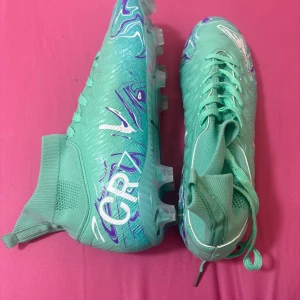 Mintgröna CR7 fotbollsskor  - Säljer ett par mintgröna  CR7 fotbollsskor med högt skaft och snörning. Skorna har ett coolt abstrakt mönster i lila, vitt och rosa samt CR7-logga på sidan. Ovandelen är i syntetmaterial med mesh vid ankeln för extra komfort. Sulan har dobbar för gräsplan.