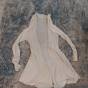 Ljusbeige stickad långklänning - Säljer en ljusbeige stickad långklänning med långärmade ärmar och snygga ribbade detaljer. Klänningen har ett öppet framstycke och ett mönstrat parti upptill som ger en cool och modern vibe. Perfekt för dig som gillar mjuka material och stilren design.