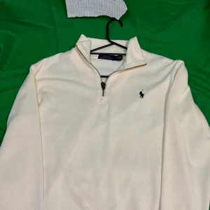 Vit half zip tröja från Polo Ralph Lauren - Vit långärmad tröja från Polo Ralph Lauren med klassisk half zip-dragkedja och broderad marinblå logga på bröstet. Tröjan har en hög krage och är tillverkad i mjuk bomull, perfekt för lager-på-lager-stil.Liknande modell som originalet!