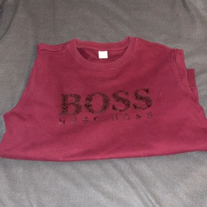 Vinröd BOSS sweatshirt med paljetter - Vinröd sweatshirt från Hugo Boss med glittrande paljettdetaljer på bröstet där det står BOSS. Tröjan har rund halsringning och långa ärmar. Perfekt för dig som vill sticka ut lite extra med en snygg logga framtill.