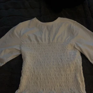 Vit smockad topp från H&M  - Säljer en vit topp från H&M  i storlek M. Toppen har smockad front och baksida som ger en snygg struktur och sitter skönt. Långa ärmar och rund halsringning. Perfekt att styla med jeans eller kjol för en trendig look.