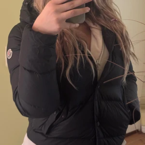 Svart Moncler pufferjacka med huva - Säljer en svart pufferjacka från Moncler med huva och diskret logga på ärmen. Jackan är quiltad och har dragkedja framtill samt knappar vid fickorna. Perfekt för kalla dagar och riktigt snygg till streetwear-stilen. Pris går att diskutera!