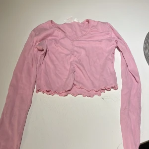 Rosa croppad topp från Gina Tricot - Söt rosa croppad topp från Gina Tricot med långa ärmar och ribbad struktur. Toppen har v-ringning, rynkdetalj framtill och vågig volangkant nertill. Perfekt för dig som gillar en trendig och feminin stil.