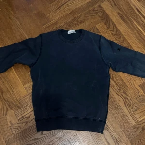 Svart sweatshirt från Stone Island Junior - Mörkblå sweatshirt från Stone Island Junior med rund hals och klassisk passform. Tröjan har långa ärmar och detaljerade knappar på ena ärmen. Tillverkad i mjuk bomull, perfekt för en clean och stilren look. Tröjan saknar dock patch vilket sänker priset.