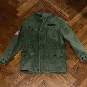 Grön parkajacka Polo Ralph Lauren - Snygg grön parkajacka från Polo Ralph Lauren med fyra stora fickor framtill, hög krage och patch med amerikansk flagga på ärmen. Jackan har röd- och svart-rutigt innerfoder och stängs med dragkedja och knappar. Perfekt för dig som gillar utility-vibe.