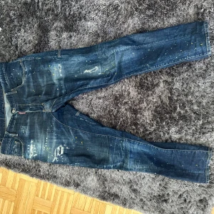 Dsquared2 blå jeans med färgstänk - Snygga blå jeans från Dsquared2 med coola slitningar och färgstänk i vitt och gult. Jeansen har klassisk femficksdesign, raka ben och tydliga sömmar. Märkeslapp bak och Dsquared2-logga vid fickan. Perfekta för dig som gillar en edgy streetstil. Köpa från farfetch för 5700kr