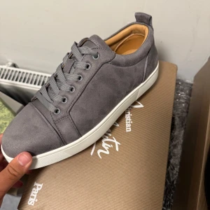 Grå mocka sneakers Christian Louboutin - Snygga grå sneakers från Christian Louboutin i mjuk mocka med klassisk röd sula och vita kanter. Skorna har rund tå, snörning och beige skinnfoder. Perfekt för dig som vill ha en clean och lyxig look med en ikonisk touch.