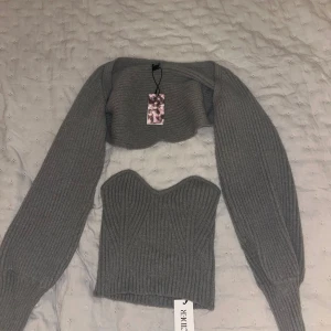 Grå stickad korsettopp med bolero från Cider - Säljer en grå stickad korsettopp med matchande croppad bolero från Cider. Toppen har en snygg sweetheart-urringning och ribbat mönster, medan boleron har långa ärmar och samma ribbade struktur. Perfekt för lager-på-lager och trendiga outfits.