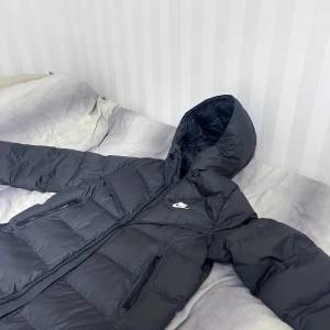 Svart lång pufferjacka från Nike - Säljer en svart lång pufferjacka från Nike med huva och två dragkedjeförsedda fickor framtill. Jackan har en tydlig quiltad design och Nike-logga på bröstet. Perfekt för kalla dagar när du vill hålla dig varm och stilsäker.