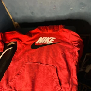 Röd Nike hoodie  - Snygg röd hoodie från Nike med stor vit och svart logga på bröstet. Klassisk känguruficka framtill och huva. Perfekt för dig som gillar sportig och avslappnad stil.