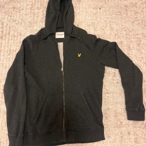 Mörkgrå hoodie från Lyle & Scott - Mörkgrå hoodie från Lyle & Scott i ekologisk bomull. Tröjan har dragkedja framtill, huva och två fickor. Klassisk gul logga på bröstet och ribbade muddar vid ärmslut och nederkant. Perfekt för chill dagar eller när du vill ha en enkel och stilren look.