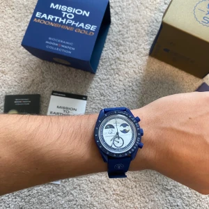 MoonSwatch Mission to Earthphase Moonshine Gold Snoopy - Omega X Swatch MoonSwatch Mission to Earthphase Moonshine Gold med blå boett och blått band. Klockan har vit urtavla med blå detaljer och kronograf-funktion. Biokeramisk material, coolt motiv på baksidan och snygga boxar med Snoopy-illustration. Perfekt för dig som gillar unika och trendiga klockor. Helt ny med allt inkluderat. Det nyaste och mest eftertraktade droppet som Omega X Swatch har gjort hittils.