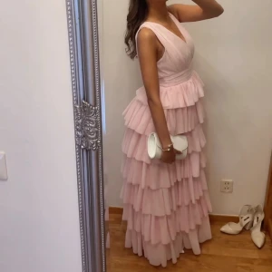 Ljusrosa volangklänning med v-ringning - Supersöt långklänning i ljusrosa med flera lager volanger och v-ringad omlott-topp. Klänningen är ärmlös och har markerad midja, perfekt för dig som vill sticka ut med en romantisk look. Materialet känns lätt och flowy, perfekt för speciella tillfällen. Endast använd 1 gg, köptes för 1800kr. Är 166 å har 5cm klackskor för referens:)🩷 Pris kan diskuteras.