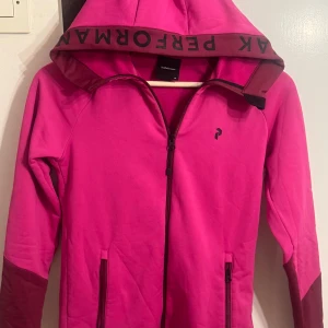 Rosa vindjacka från Peak Performance - Snygg rosa vindjacka/kofta från Peak Performance med huva och dragkedja framtill. Jackan har mörkare detaljer på ärmslut och huva, samt två fickor med dragkedja. 