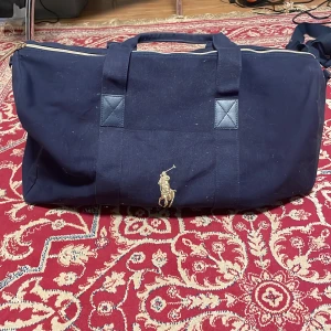 Duffel från Polo Ralph Lauren - Snygg mörkblå duffel från Polo Ralph Lauren med klassisk guld broderad polospelare på framsidan. Väskan har robusta handtag, dragkedja i guld och är tillverkad i kraftig bomullscanvas. Perfekt för resor eller gymmet.