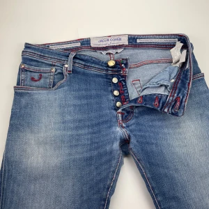 Jacob Cohën jeans 622 - Snygga blå jeans från Jacob Cohën med röda kontrastsömmar och en röd patch bak. Jeansen har klassisk femficksdesign, knäppning med knappar och cool faded effekt. Perfekta för dig som gillar detaljer och vill sticka ut lite extra.