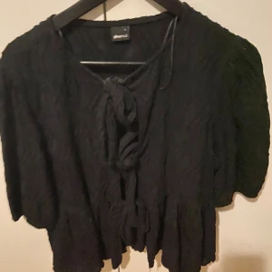 Svart blus med knyt från Gina Tricot - Svart blus från Gina Tricot i storlek s. Blusen har korta ärmar, strukturmönstrat tyg och flera knyt framtill. Modellen är croppad och har en luftig känsla, perfekt att styla med höga jeans eller kjol.
