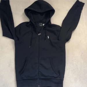Svart hoodie från Polo Ralph Lauren XS - Svart hoodie från Polo Ralph Lauren i storlek XS. Klassisk design med dragkedja, huva med snörning och två fickor framtill. Liten vit broderad logga på bröstet. Perfekt för en chill och stilren look. Nypris 2200kr köpt på zalando,  Har nog kvitto på mail om de behövs.Passar folk som är från 165-175 cm långa, Använd MAX 5 gånger om ens det! Precis som ny