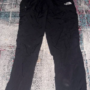 Svarta täckbyxor från The North Face - Svarta täckbyxor från The North Face med raka ben och diskret logga på höften. Byxorna har praktiska fickor och är tillverkade i ett slitstarkt syntetmaterial som passar perfekt för kalla dagar utomhus.