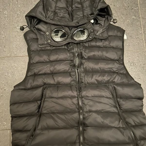 Svart dunväst från C.P. Company - Säljer en svart dunväst från C.P. Company med ikonisk huva med inbyggda goggles. Västen har quiltad design, två fickor med dragkedja och en snygg, sportig look. Perfekt för lager-på-lager och streetwear-stil. Var för stor för mig, passar som M